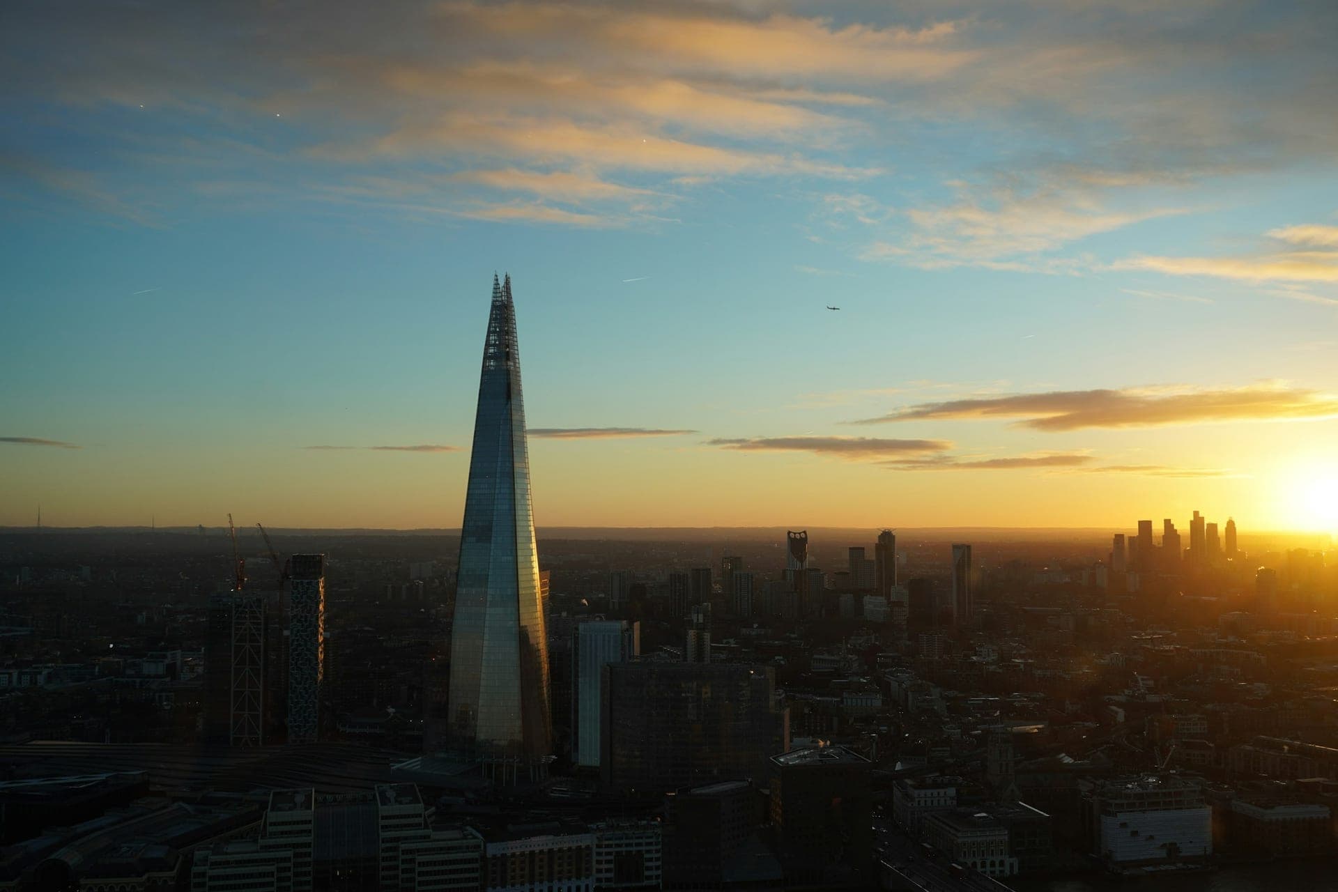 The Shard rising over London at sunset, the City skyline and Thames basin glowing gold under a calm blue-and-amber dusk sky