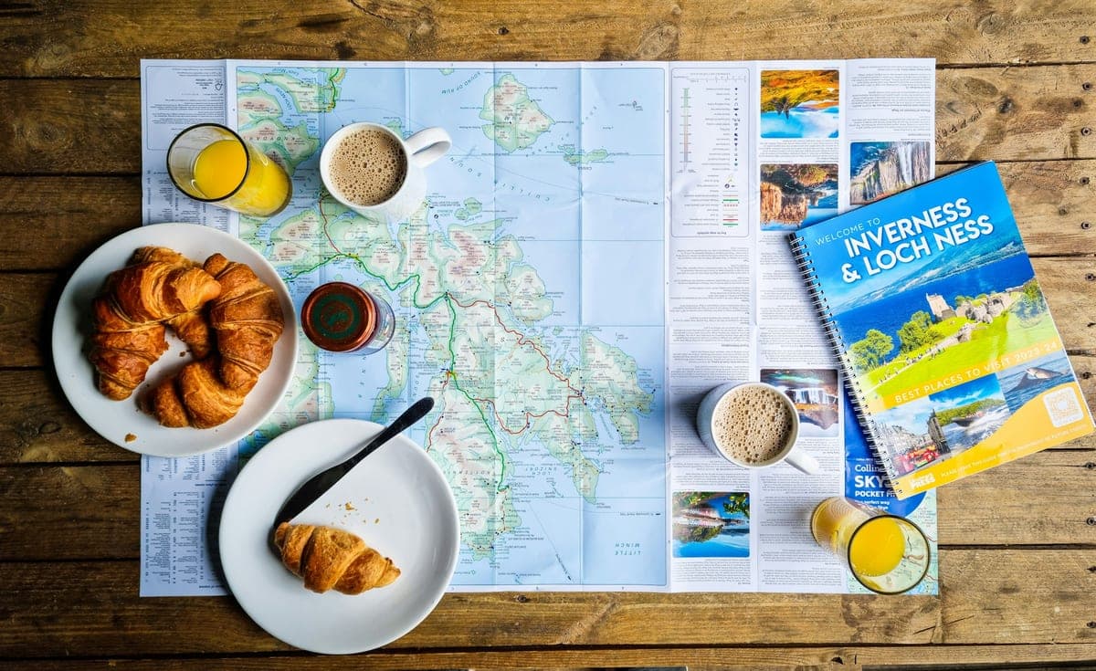 A travel map spread on a table with fresh croissants, a glass of orange juice and a warm morning light — the relaxed feel of planning an outing