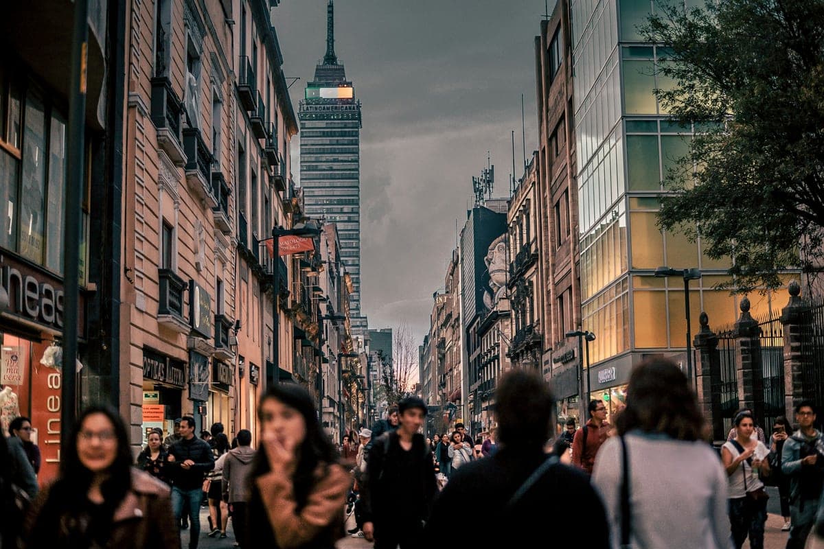 Real people walking a warm, lamplit city street at dusk, shop windows glowing under a moody evening sky — the after-work city coming alive