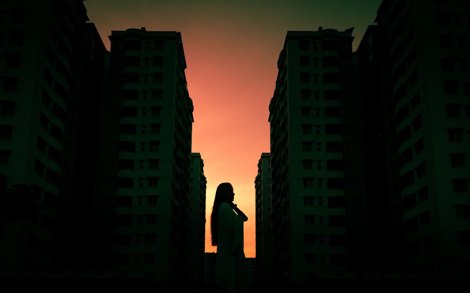 A lone figure in silhouette standing in the gap between two tall city towers, backlit by a blazing orange-and-amber sunset — solitary, but warm