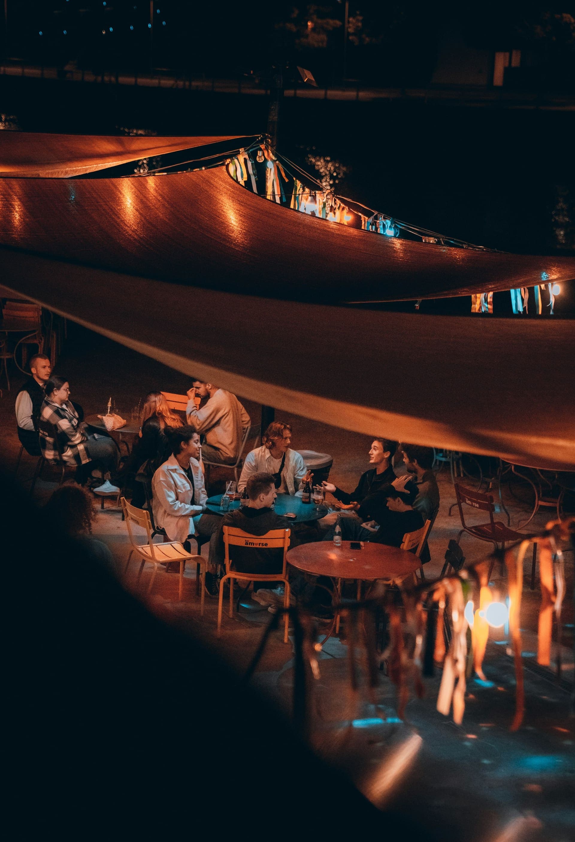 A candid group of friends gathered around tables under warm golden string lights at an outdoor venue at night, glasses on the table, relaxed after-work evening mood