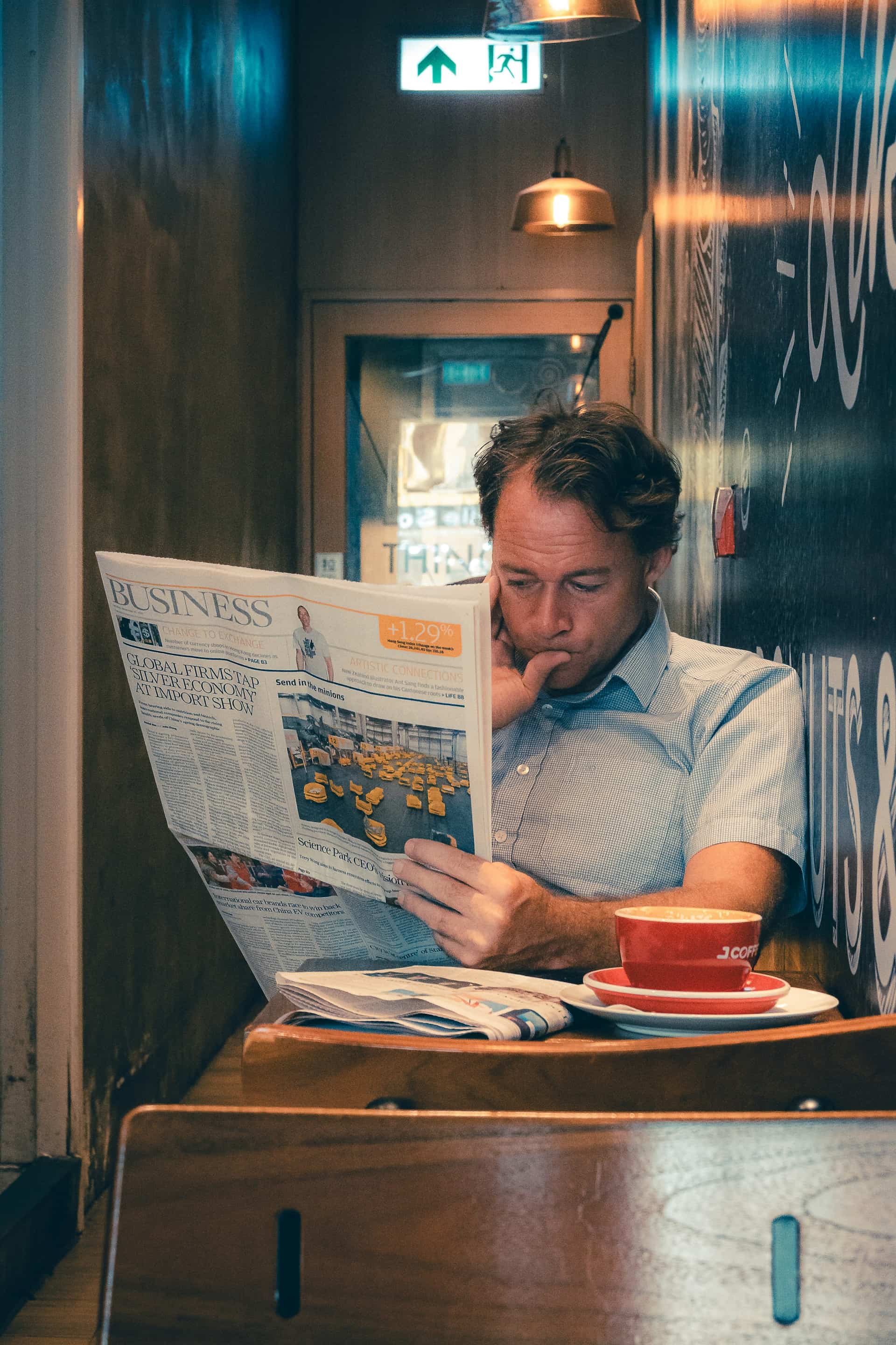 A man reading a printed BUSINESS broadsheet over a flat white in a warm, dimly-lit London bar at night, golden tungsten light, real candid evening scene