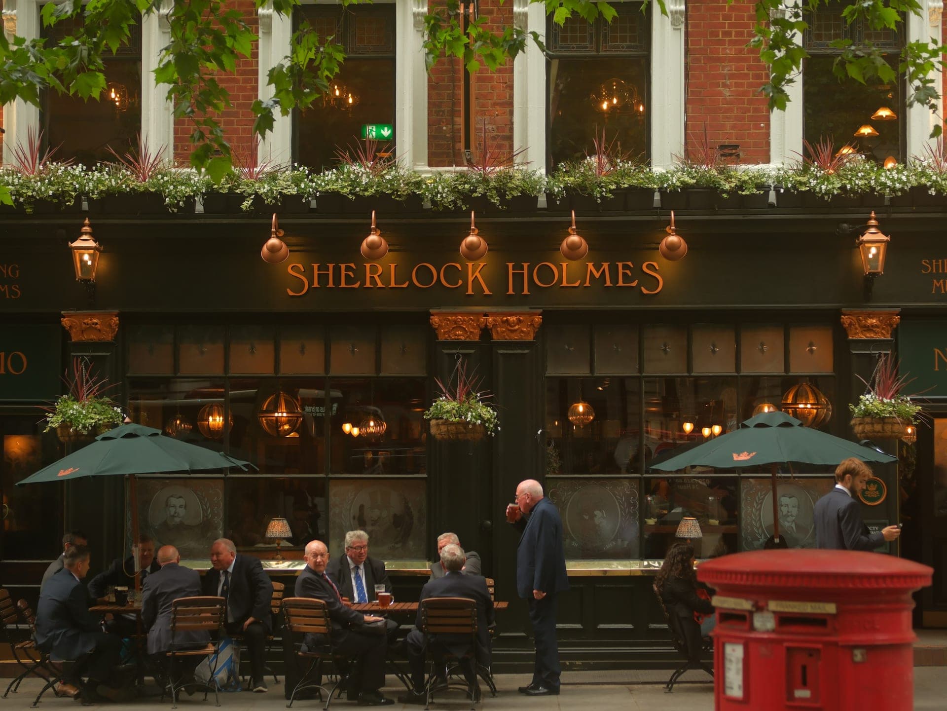 Real people drinking at outdoor tables outside the characterful green-and-copper Sherlock Holmes pub in London on a warm evening, plane-tree leaves above and a red postbox in the foreground