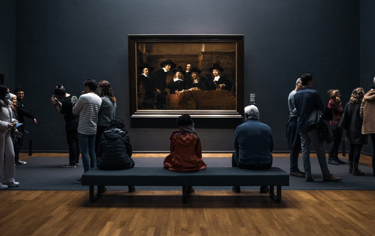 Visitors gathered in a dimly lit museum gallery in the evening, some seated on a bench, contemplating a large old-master painting under warm light against dark walls