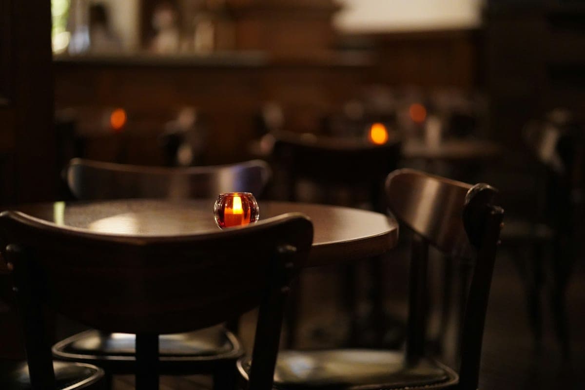 A single amber candle glowing on a bare wooden table in a warm, dimly-lit characterful restaurant at dusk, empty bentwood chairs around it