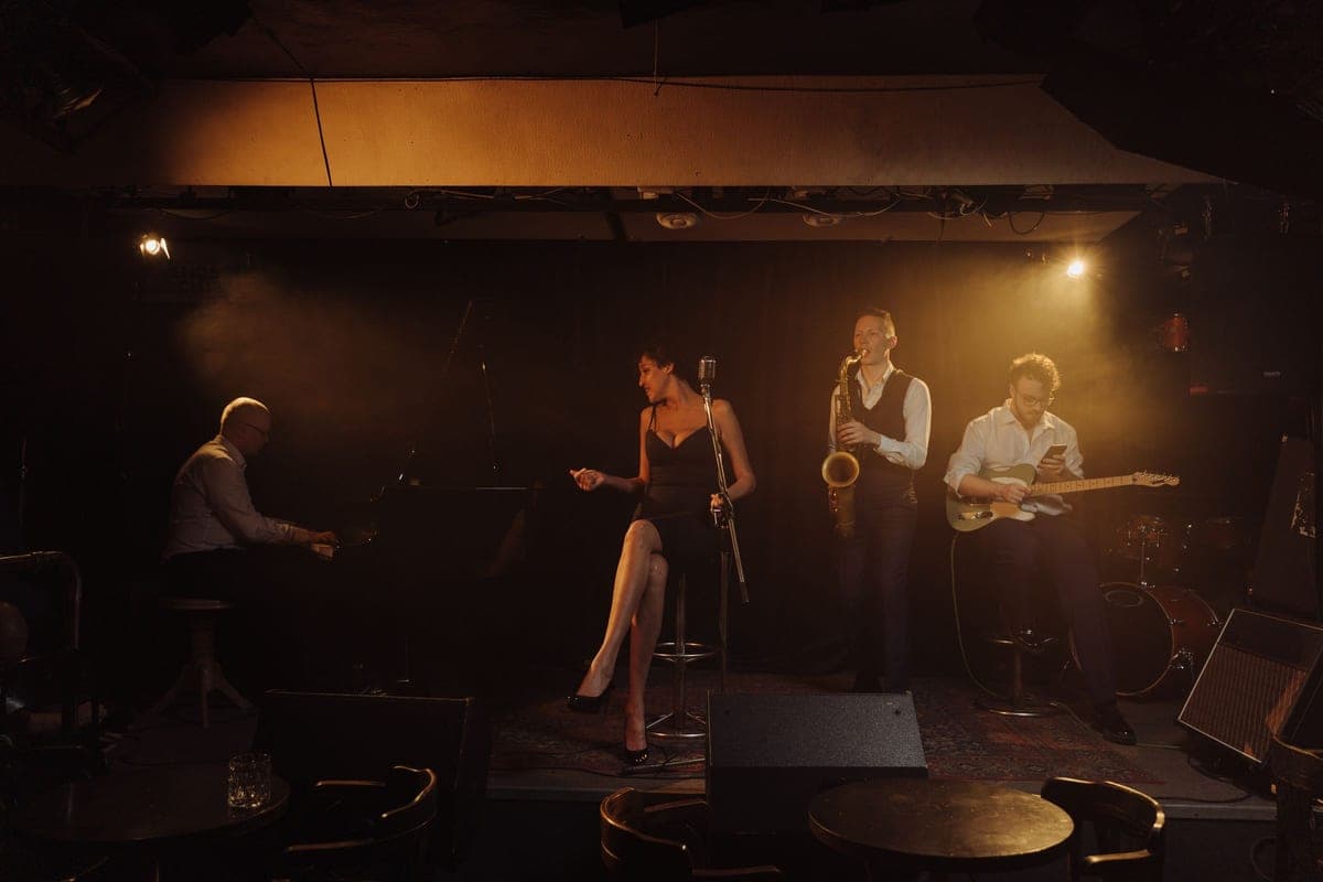 A jazz quartet performing on a small, warmly-lit club stage with dark candlelit tables in the foreground