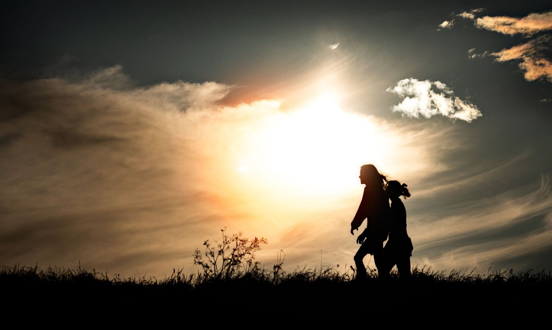 Two people walking close together in bold silhouette along a ridge, backlit by a blazing golden-hour sun through warm wispy clouds
