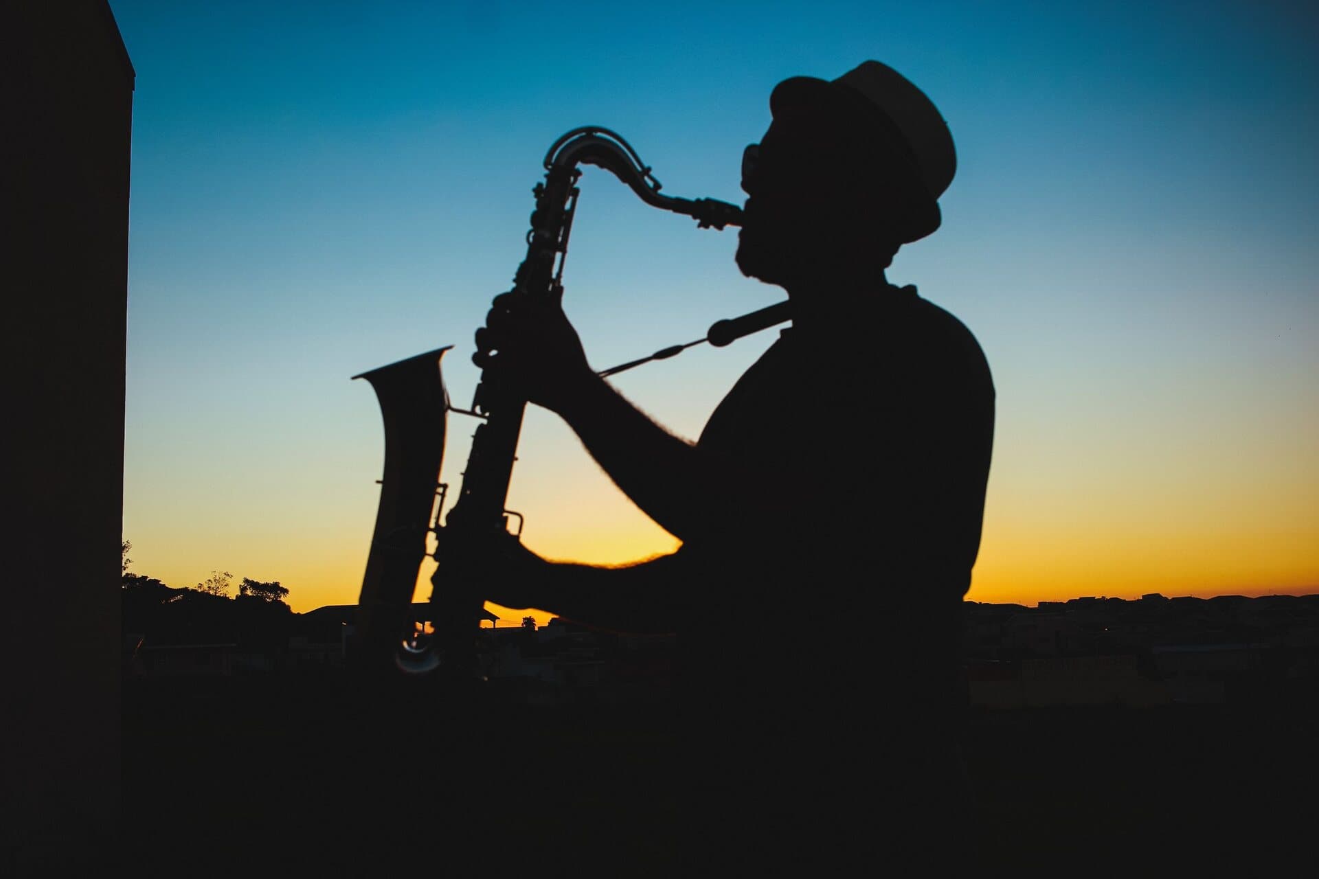 A saxophonist in a fedora in bold silhouette, playing against a glowing blue-to-amber sunset over the city