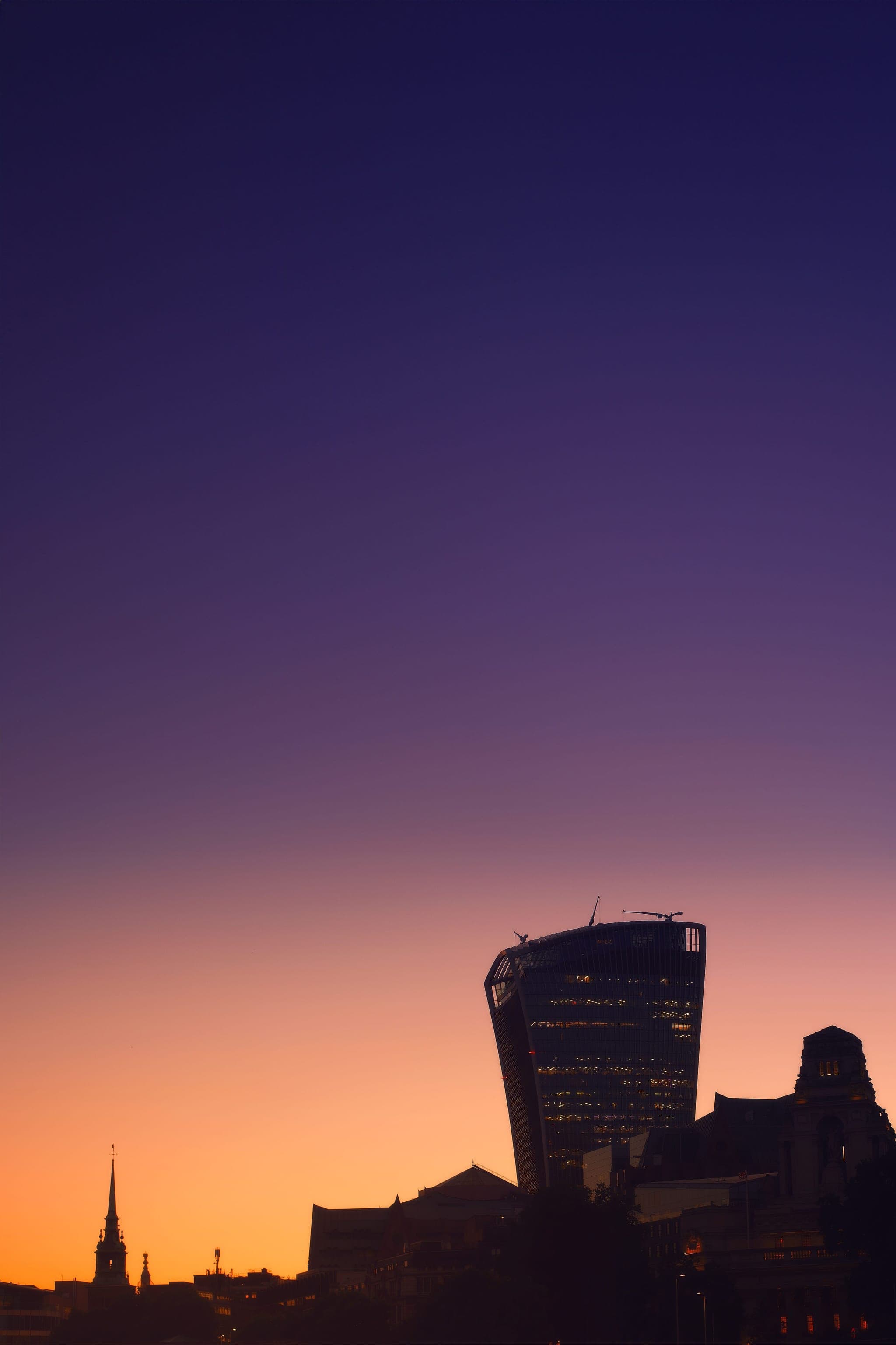 London at twilight — the City of London skyline silhouetted against a deep blue-to-purple night sky that melts into a pink-and-gold sunset glow at the horizon; the rounded Walkie-Talkie tower stands lit on the right, a church spire and riverside buildings on the left