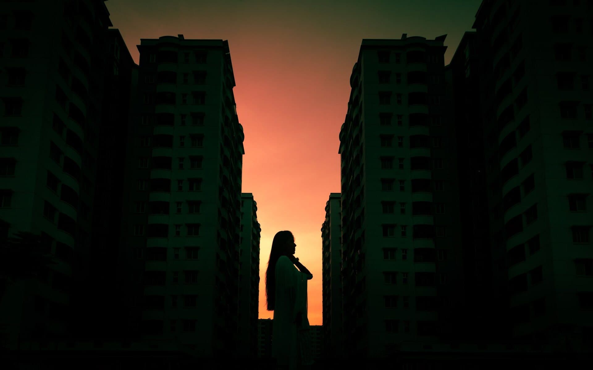 A lone figure in silhouette standing between two dark high-rise towers, backlit by a blazing orange-and-magenta sunset glowing in the gap