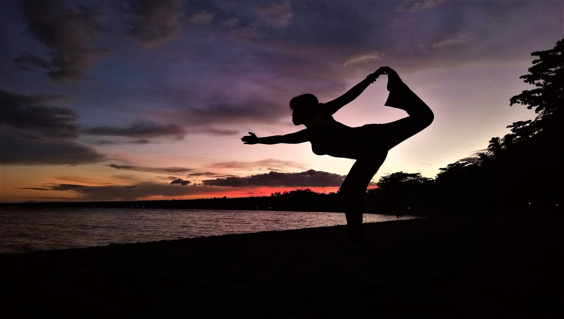 A lone figure holding a graceful yoga pose in silhouette against a deep purple-and-amber dusk sky over calm water