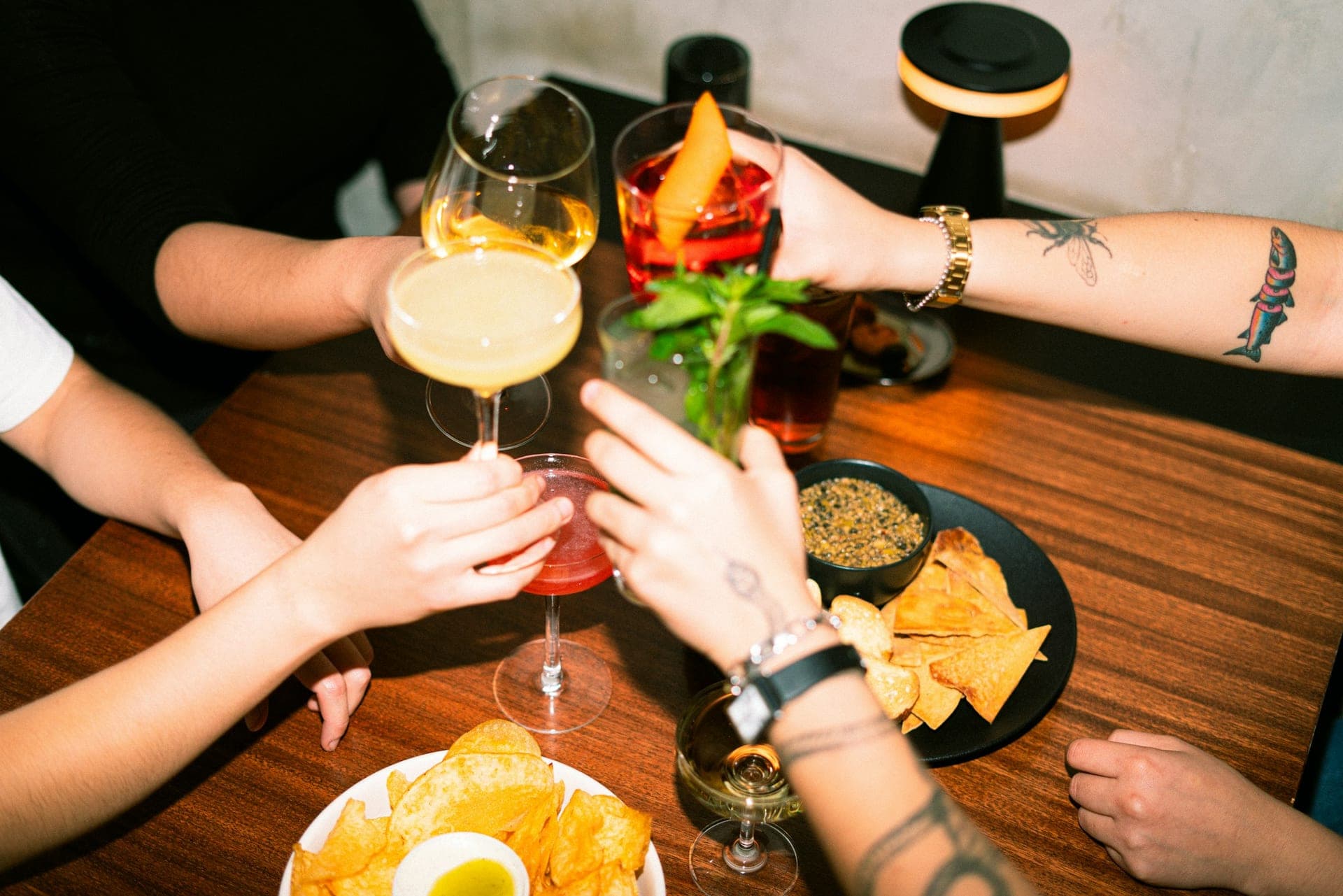 Friends raising warm-lit cocktails together over a dark wood table at a London bar after work