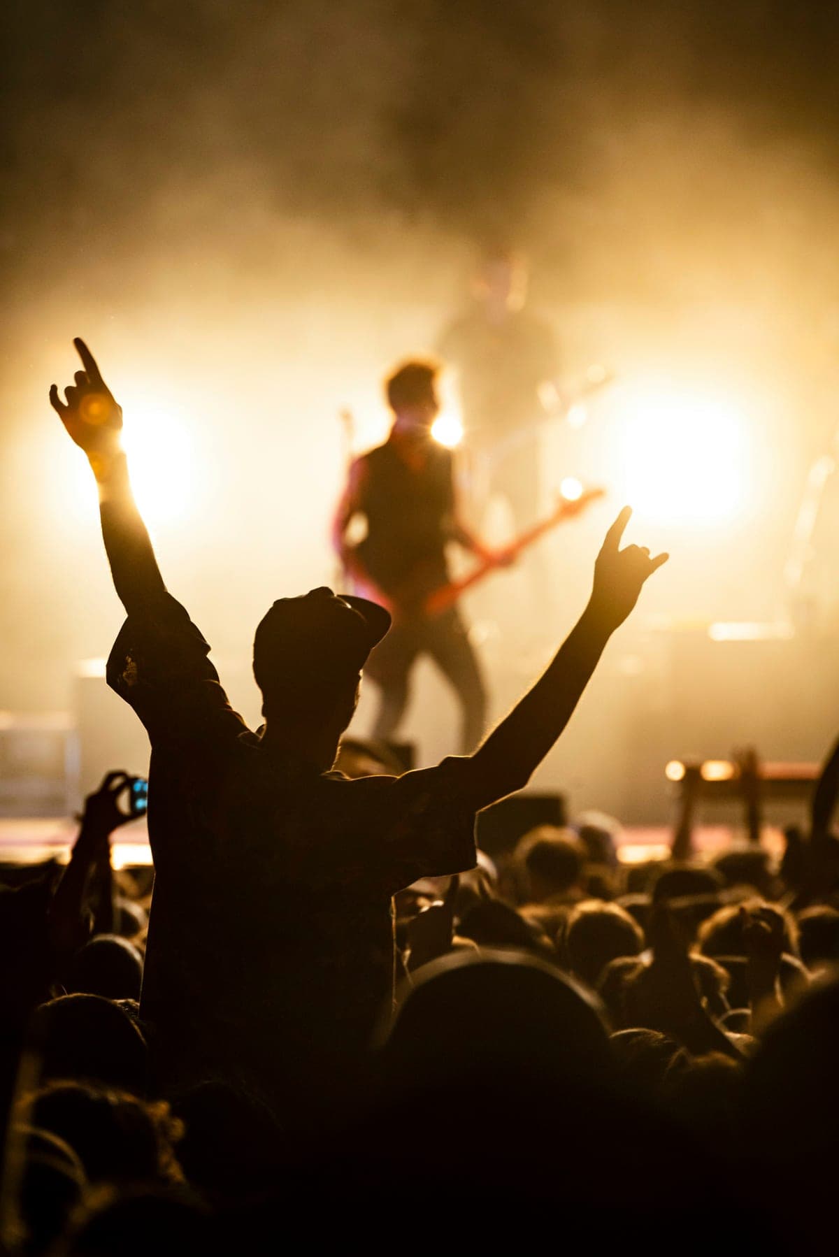 A single performer silhouetted against warm golden stage haze while a small, candid crowd raises their hands at an intimate evening gig