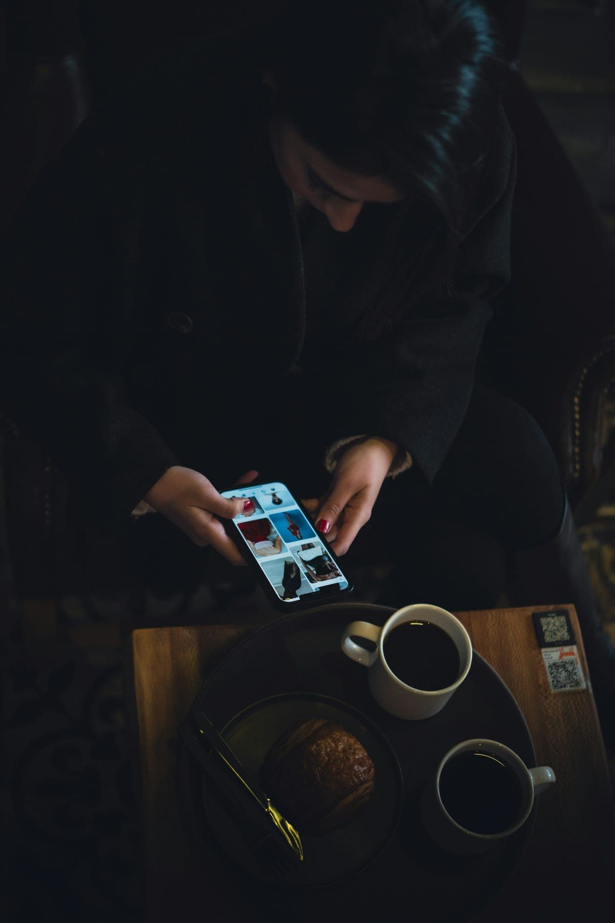Overhead candid shot of a woman scrolling her phone over coffee and a croissant at a moody, warmly-lit cafe table, browsing options for her evening