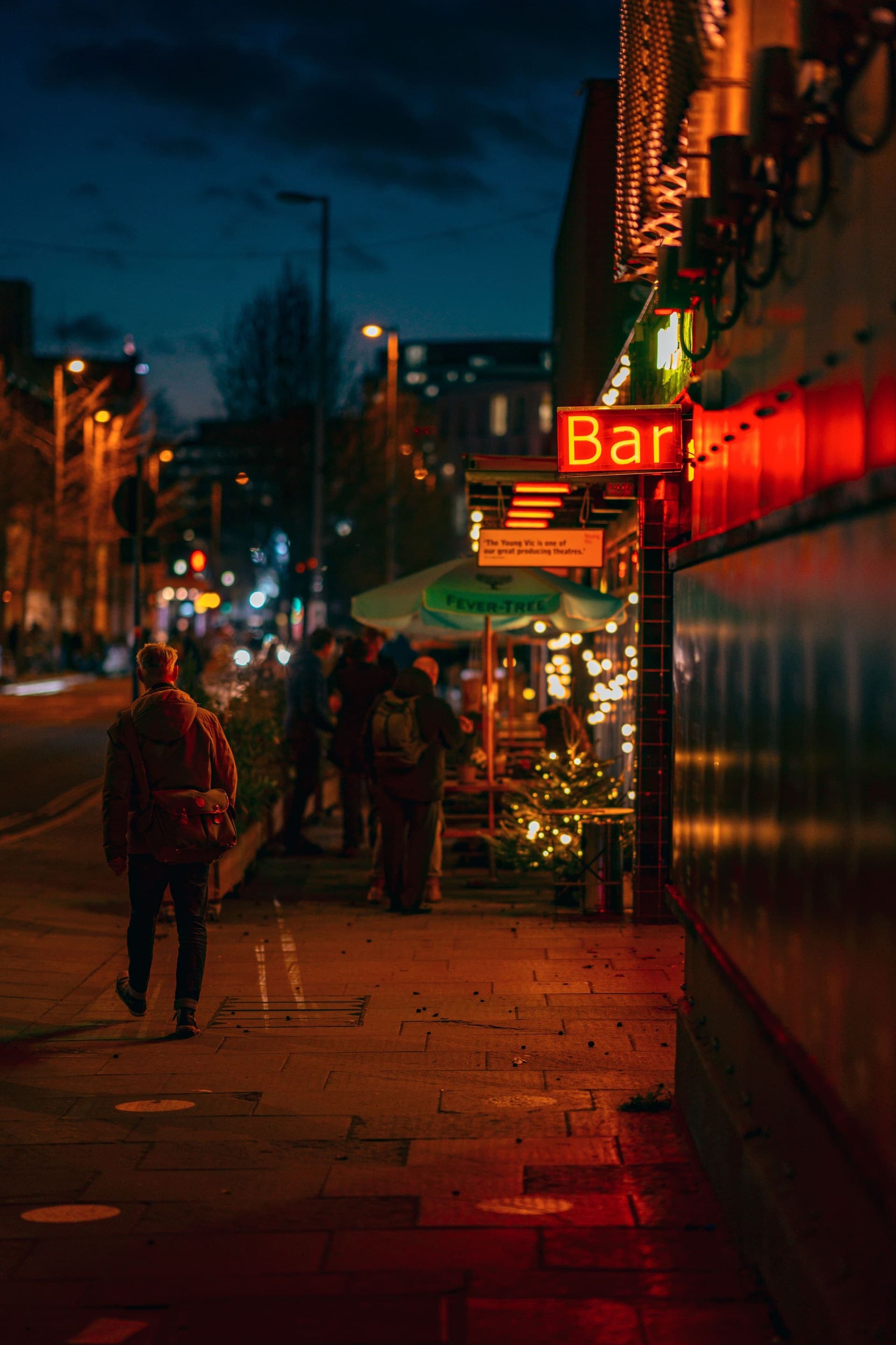 A man walks past a glowing neon "Bar" sign on a London pavement at blue hour, people gathered outside the warmly lit venue under a deep dusk sky