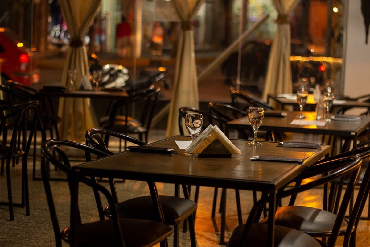 A warmly lit restaurant at dusk, its set tables and wine glasses waiting empty as people pass by on the street beyond the windows
