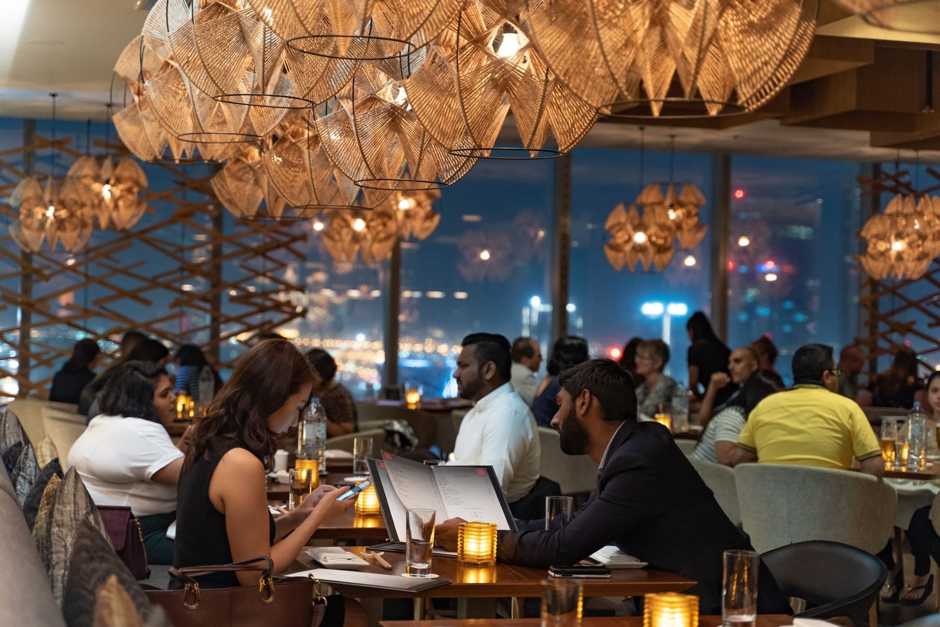 Diners at warm candlelit tables in an upper-floor restaurant at dusk, the city skyline glowing blue through floor-to-ceiling windows behind them