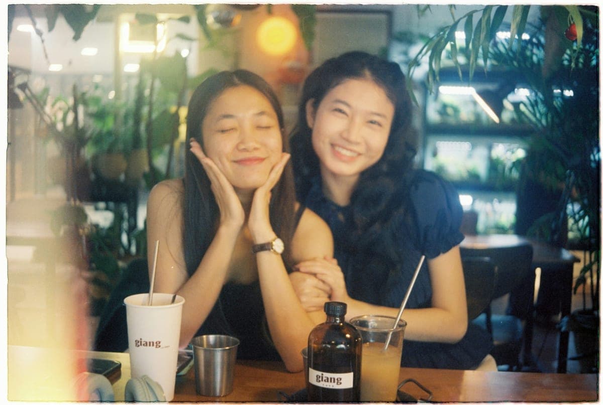 Two friends sitting close together at a warm, plant-filled cafe table in the evening, smiling over their drinks — a candid, intimate moment captured on film.