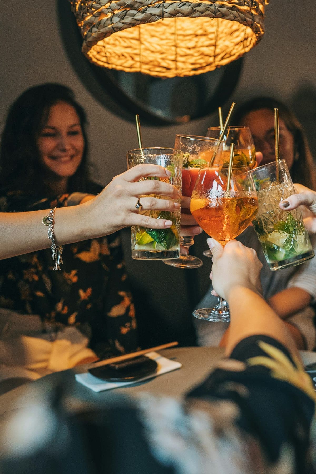 Friends clink fruit-garnished cocktails across a low table, lit by the warm glow of a woven rattan pendant lamp in a cosy bar